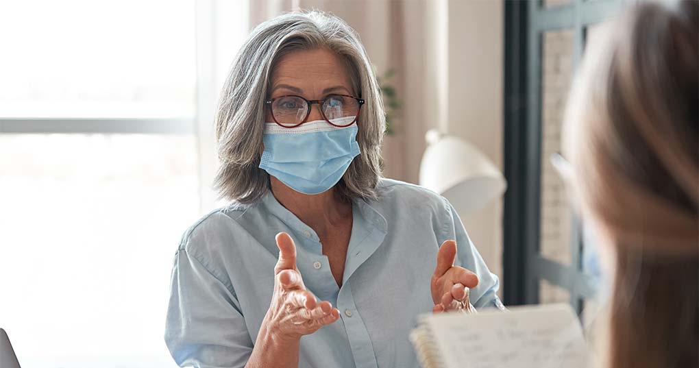A woman wearing a mask sitting across from someone taking notes