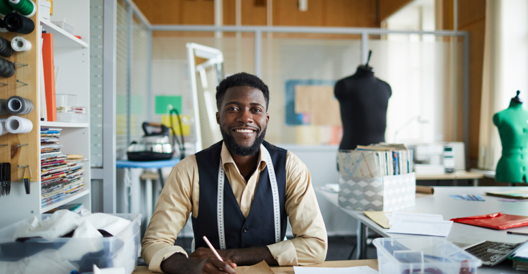 photo of smiling man in yellow shirt and sewing studio writing in notebook